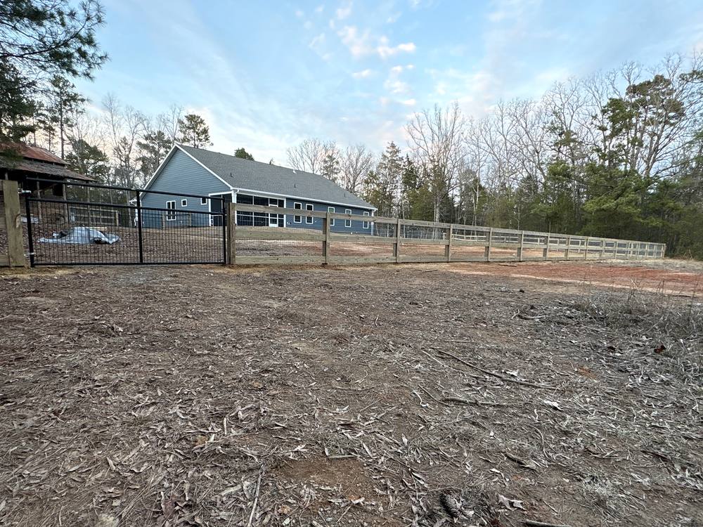 Three rail wood fence with wire mesh and a black gate in front of a blue farmhouse in the Midlands of South Carolina